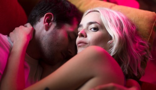 A woman and a man embrace in bed while the woman stares into the camera with a cold look.