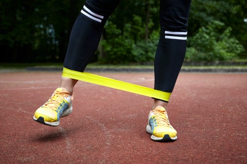 A woman's legs in exercise gear training in a park, stretching a resistance band between two feet.