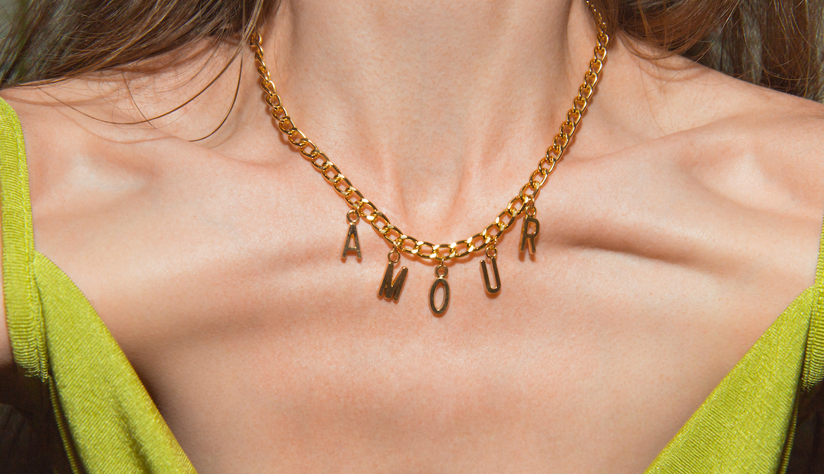 A close-up of a gold necklace around a woman's neck. The necklace contains charms that spell out the word amour, which is French for love. This photo is being used to promote an article answering the question "what is gold vermeil?"