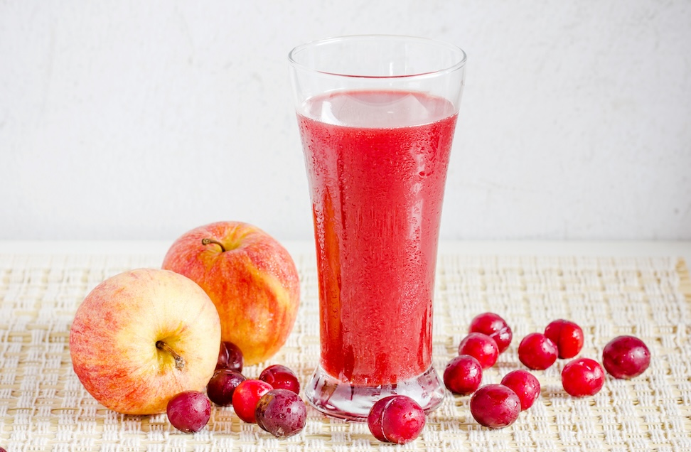 tall glass of red cranberry apple juice on placemat surrounded by whole fresh cranberries and apples