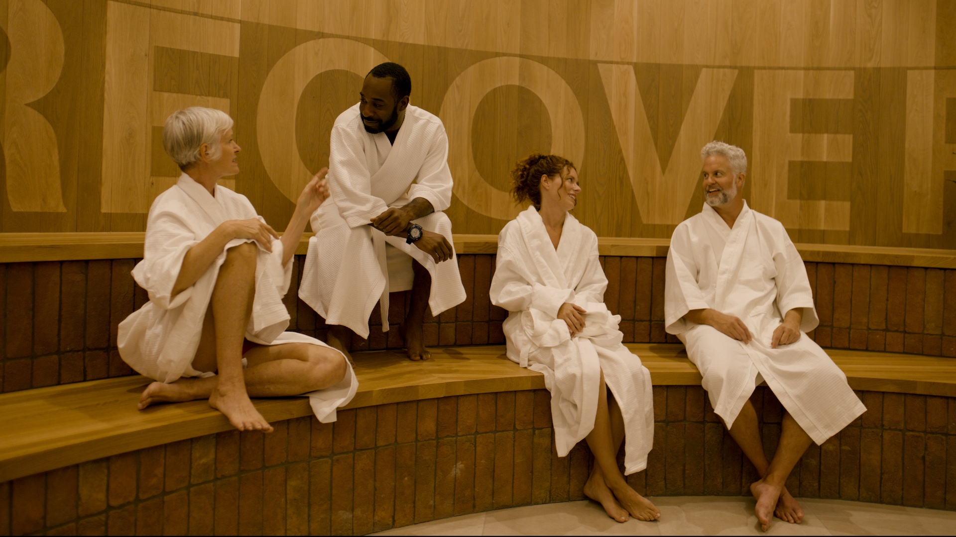 Four people lounging on wooden benches in robes with the word RECOVER on the wall behind them.