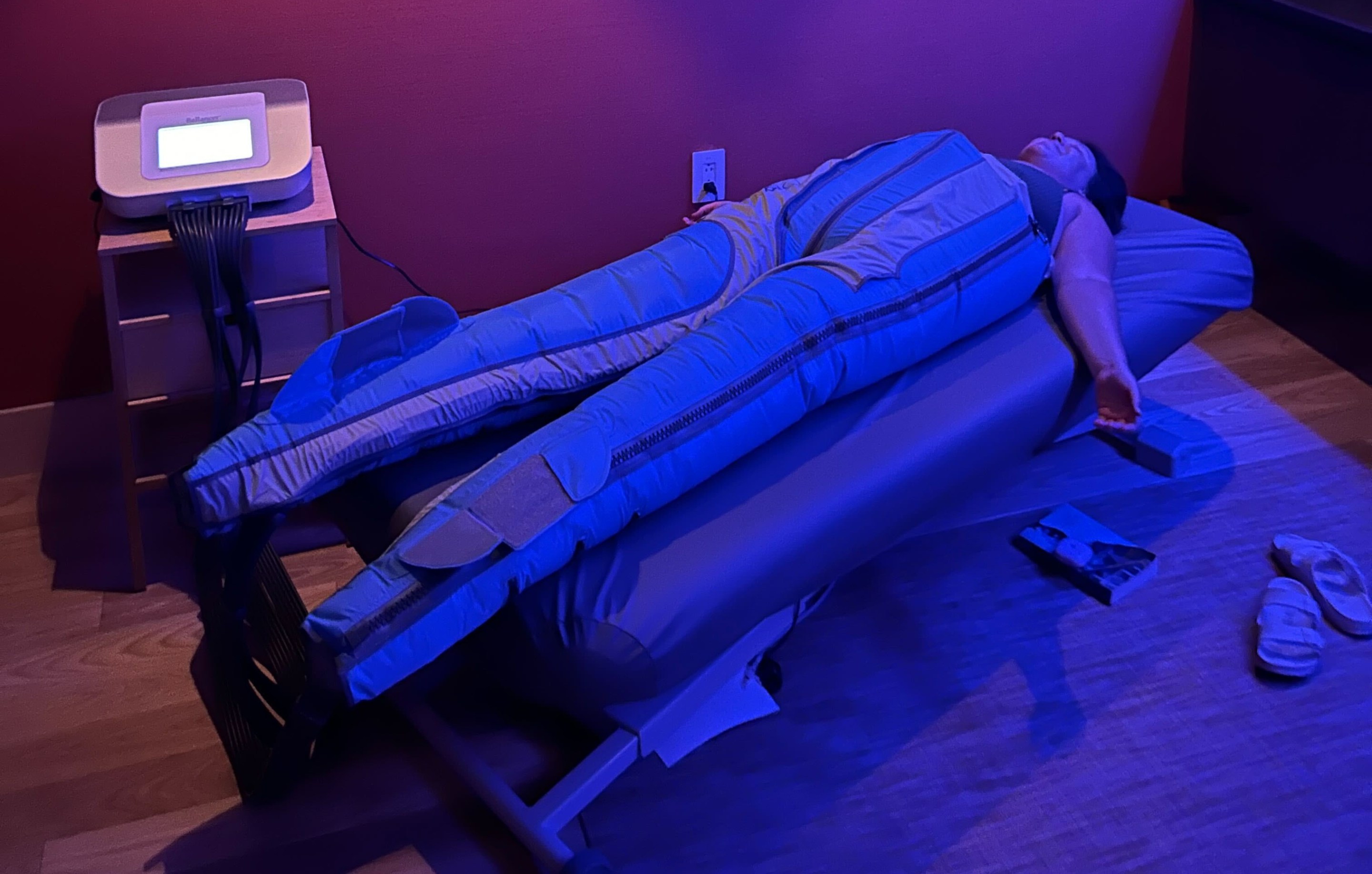 A woman (the author) laying on a bed in a full body compression suit lit by purple lighting.