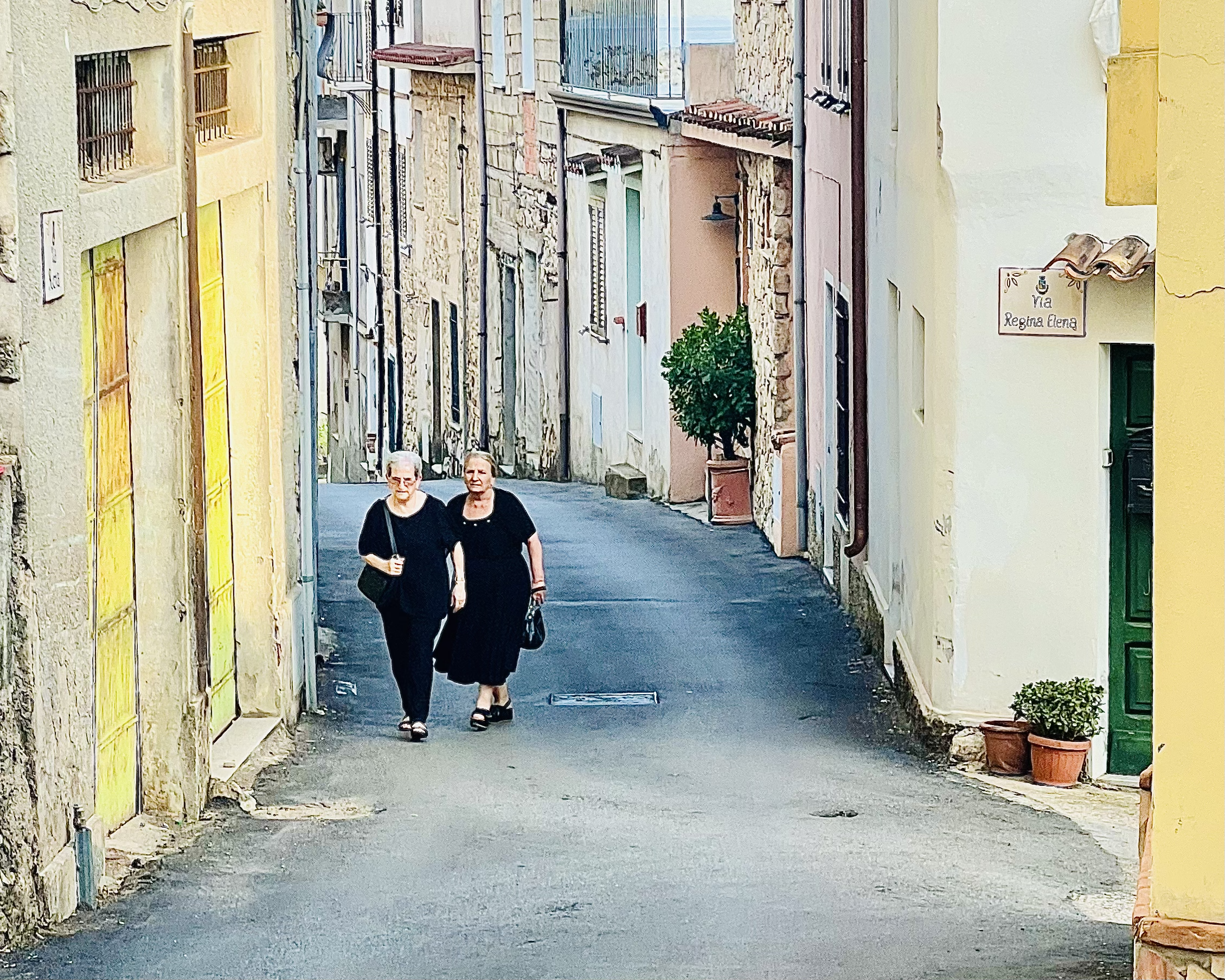 Two Sardinian women walk uphill in a narrow street in Baunei. 