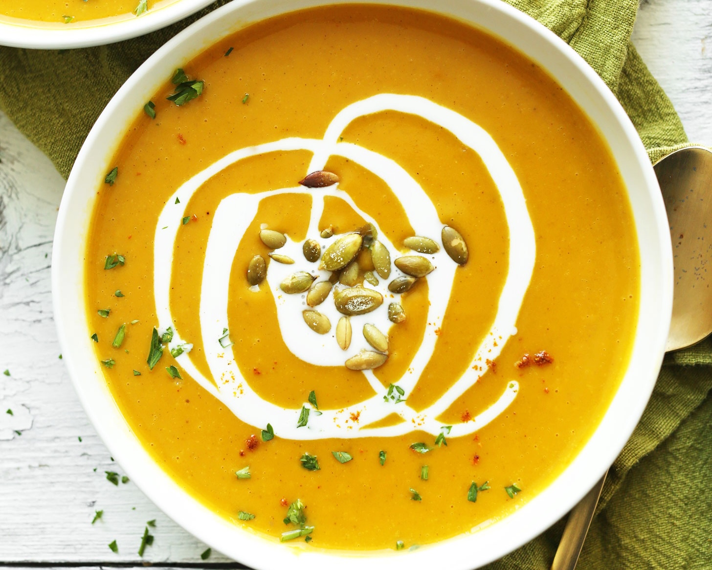 a white bowl of orange soup with a drizzle of cream and pumpkin seeds sits on a white surface