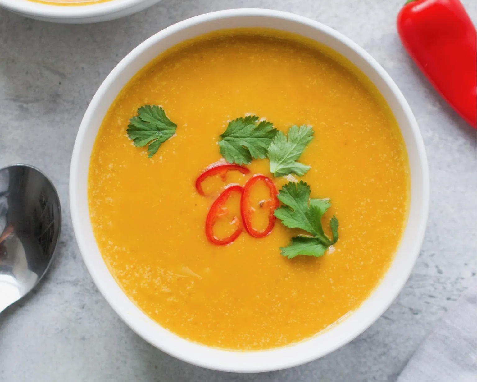 In a white bowl, orange soup garnished with green leaves and red pepper slices