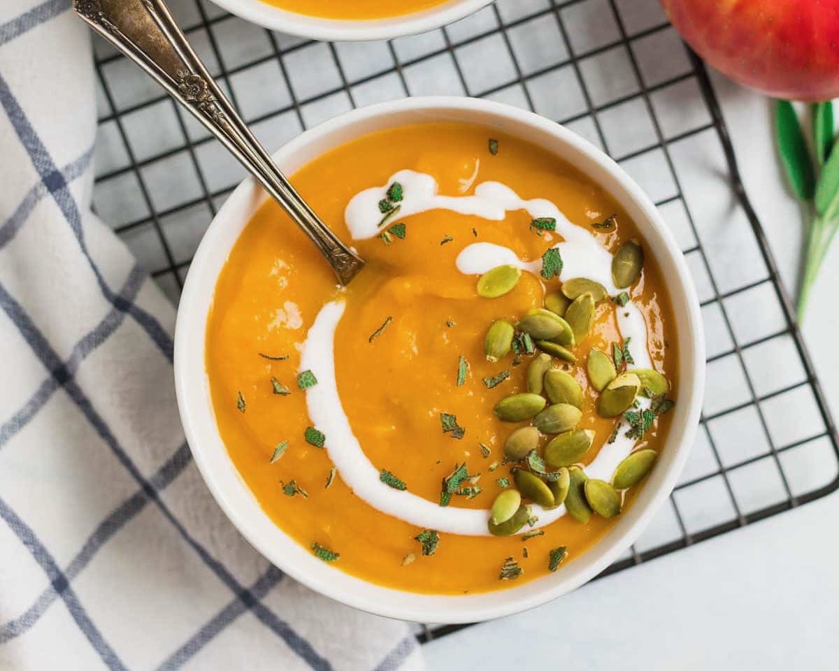 Orange soup with a drizzle of cream and pumpkin seeds rests in a white bowl on a checkered surface with a similarly textured cloth nearby