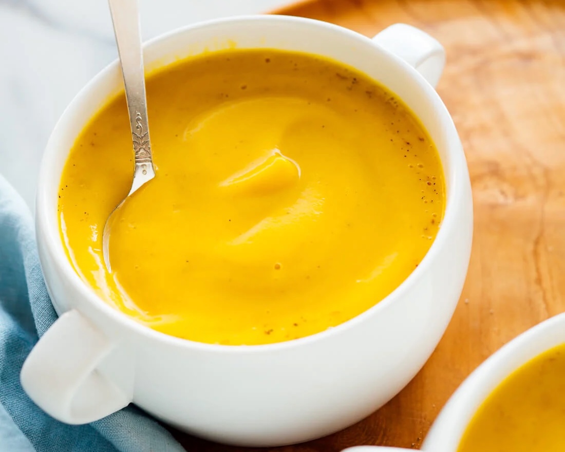 a white mug of orange soup with a spoon inside sits on a light brown surface