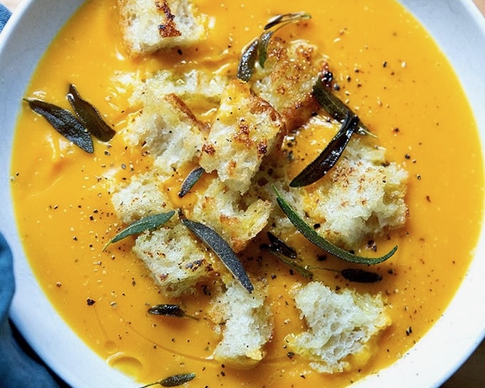 Orange soup garnished with croutons and herbs in a white bowl