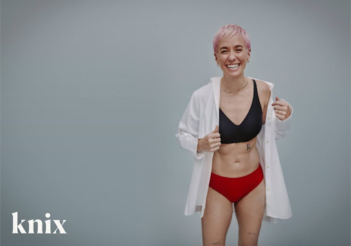 photo of megan rapinoe in black sports bra, red underwear, and white button up shirt posing for brand "knix"